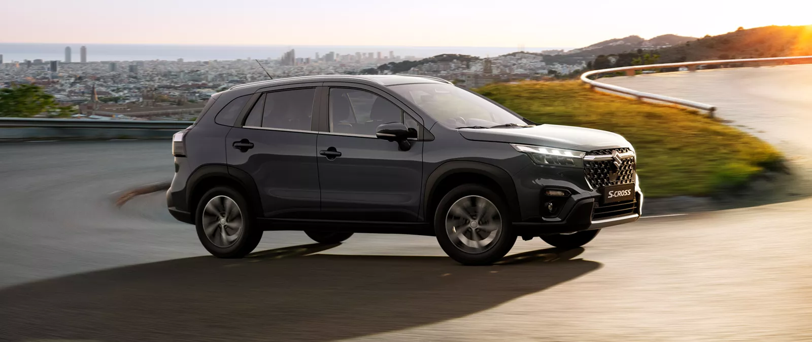 Suzuki S-Cross in Titan Dark Grey driving along a curved hillside road at sunset, with a panoramic cityscape in the background.