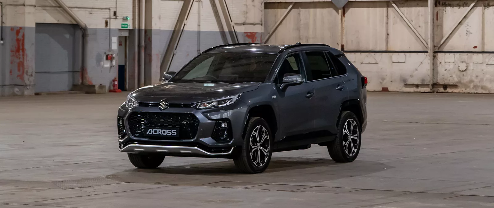 Front three-quarter view of a Suzuki Across SUV in a grey metallic finish, positioned in an industrial warehouse setting.