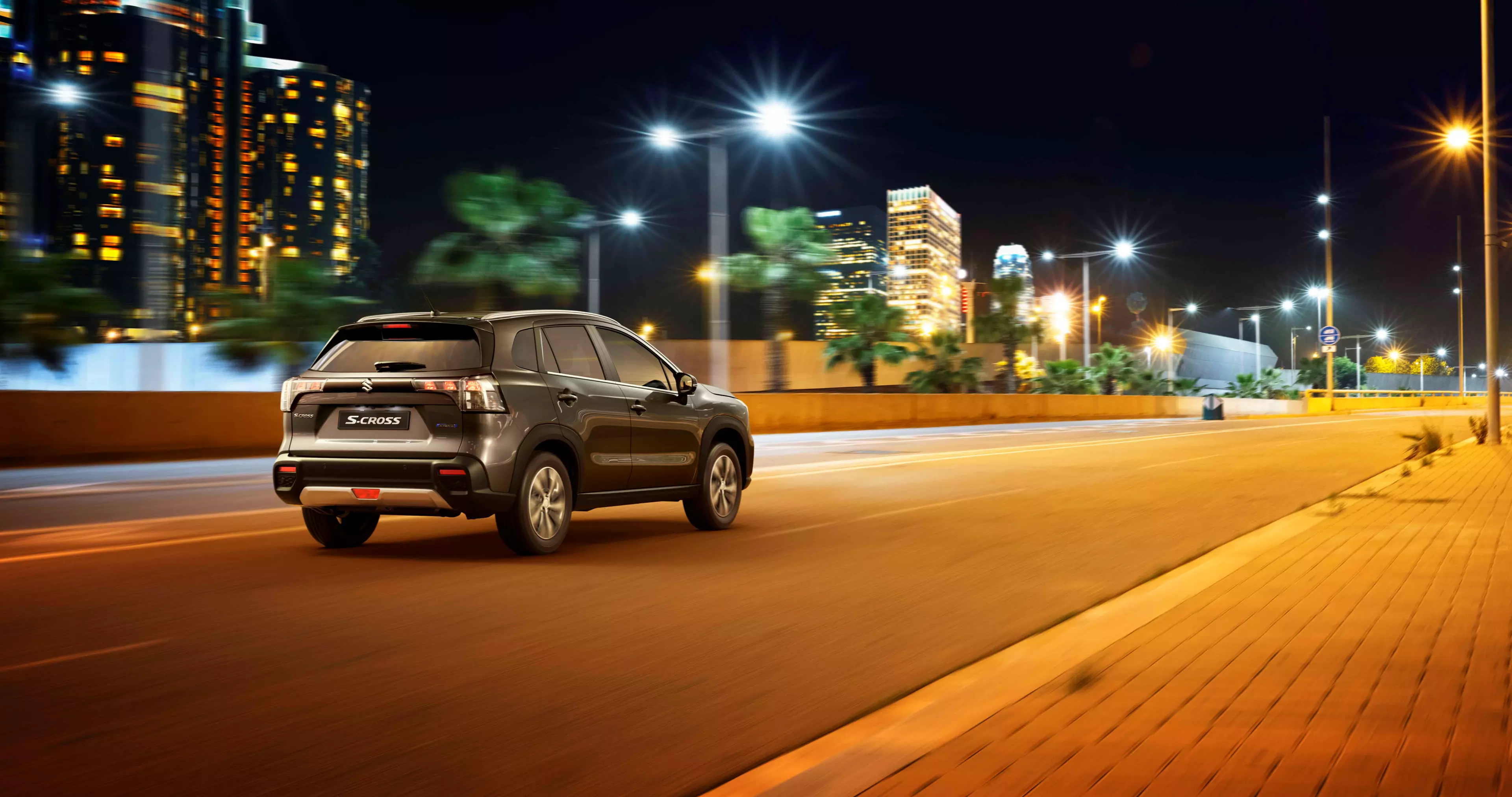 Side and rear view of a grey Suzuki S-Cross on an asphalt road in a city at night time.