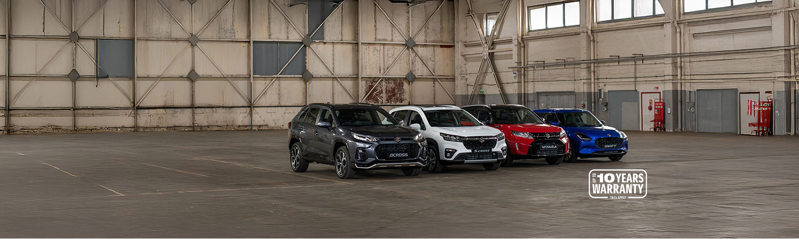 Full Suzuki car range line up including Across, S-Cross, Vitara and Swift, displayed in a large industrial warehouse with 10-year warranty badge