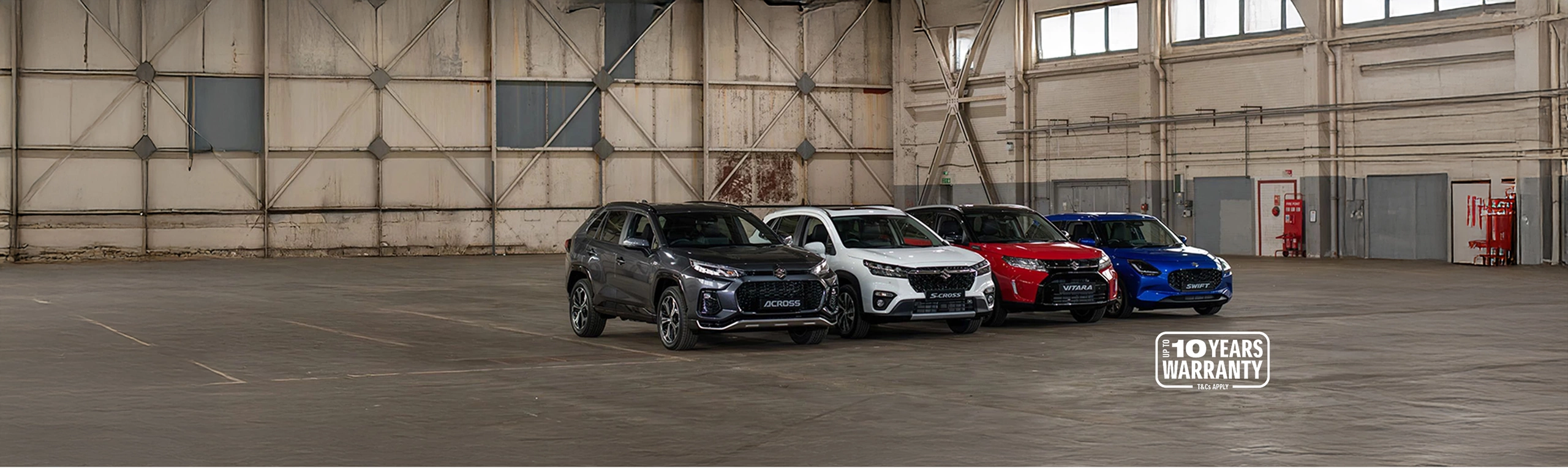 Full Suzuki car range line up including Across, S-Cross, Vitara and Swift, displayed in a large industrial warehouse with 10-year warranty badge