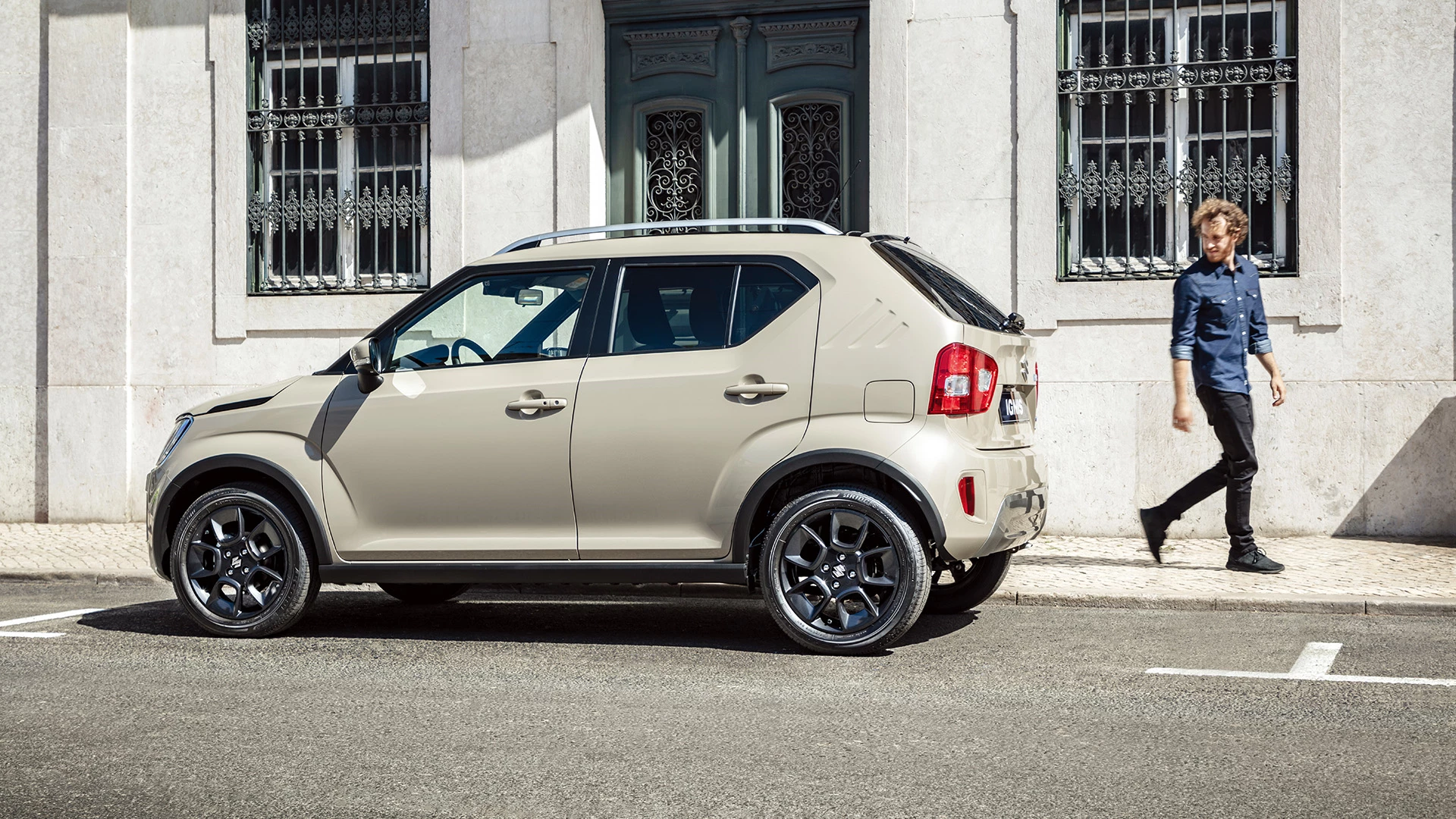 Side view of the Suzuki Ignis in Caravan Ivory parked on street, highlighting compact design and black alloy wheels.