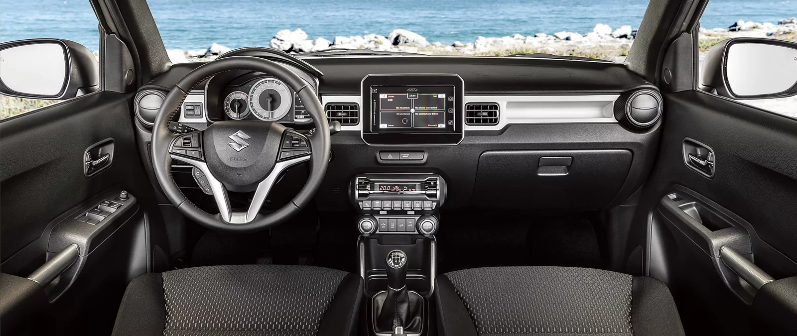 Interior view of Suzuki Igns showcasing dashboard, steering wheel and infotainment screen with ocean view in background.