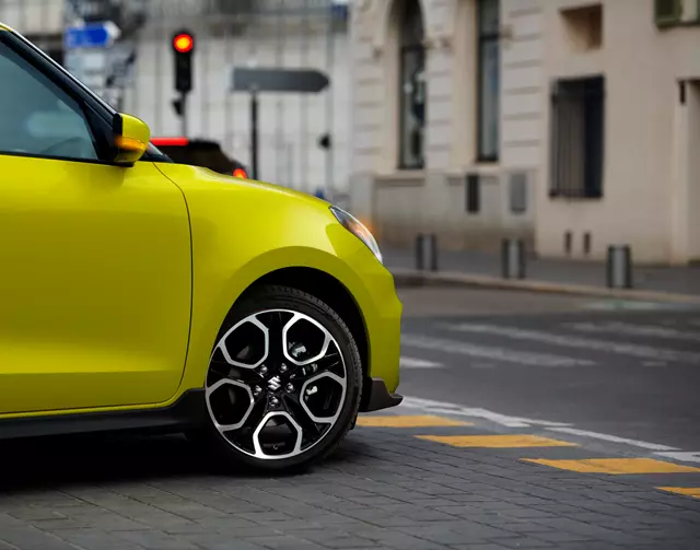 Close-up of a yellow Suzuki Swift Sport's front wheel and fender on a city street, with traffic lights and buildings in the background.