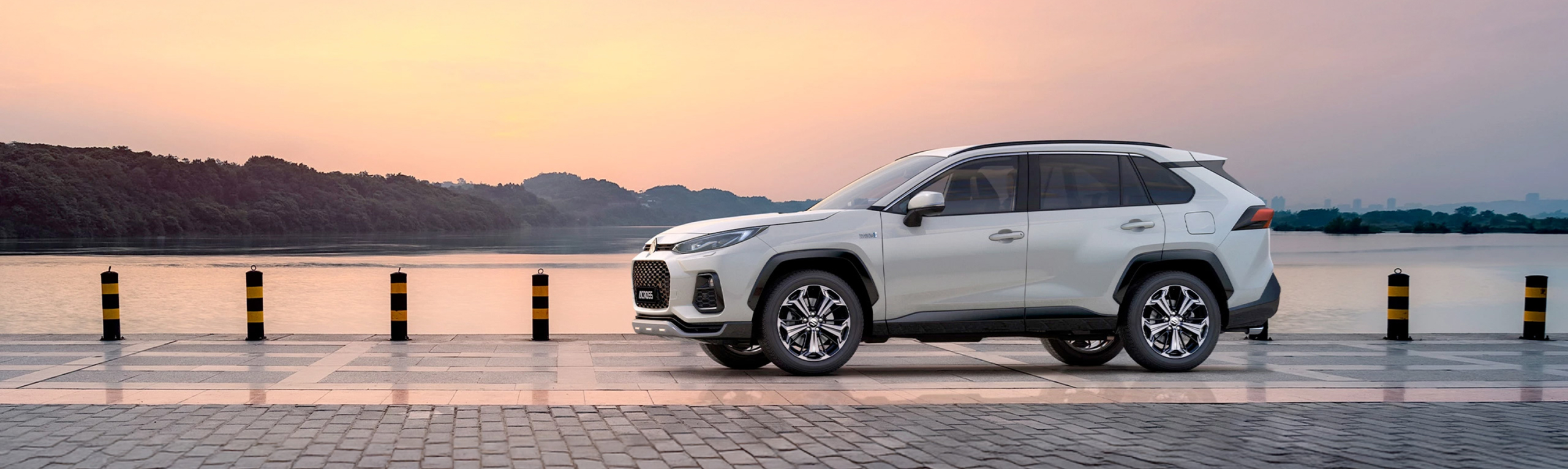 Hero banner of the Suzuki Across SUV parked on a waterfront promenade at sunset, showcasing its sleek exterior design and chrome alloy wheels.