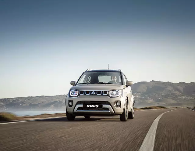 Front view of a caravan ivory metallic Suzuki Ignis on a winding coastal road with mountains in the distance.