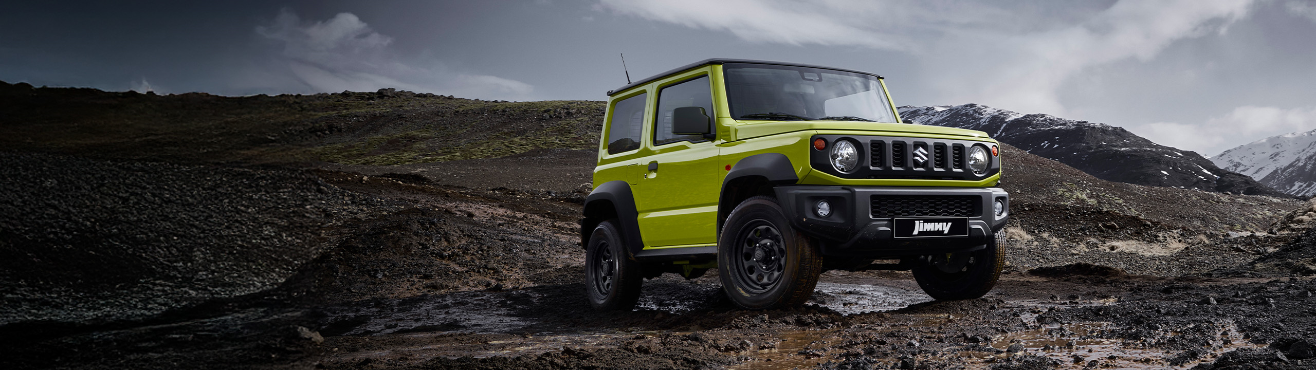 Suzuki Jimny in kinetic yellow positioned on rugged off-road terrain, front-side view with mountainous backdrop.