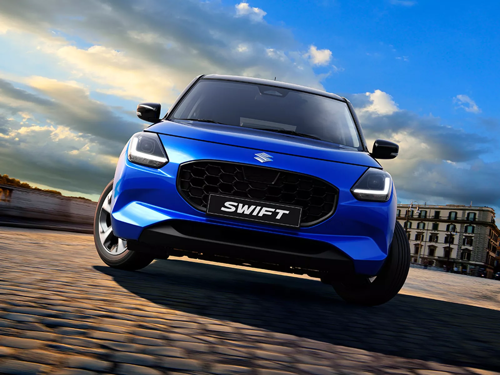 Front view of the Suzuki Swift in frontier blue, showcasing its bold grille and modern exterior design on a cobbled street backdrop.