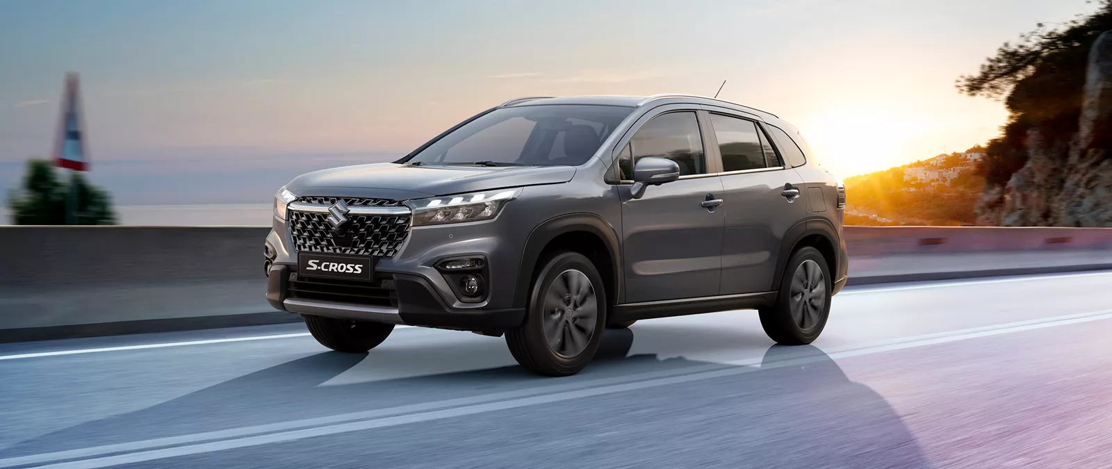 Suzuki S-Cross in Titan Dark Grey driving along a curved hillside road at sunset, with a panoramic cityscape in the background.