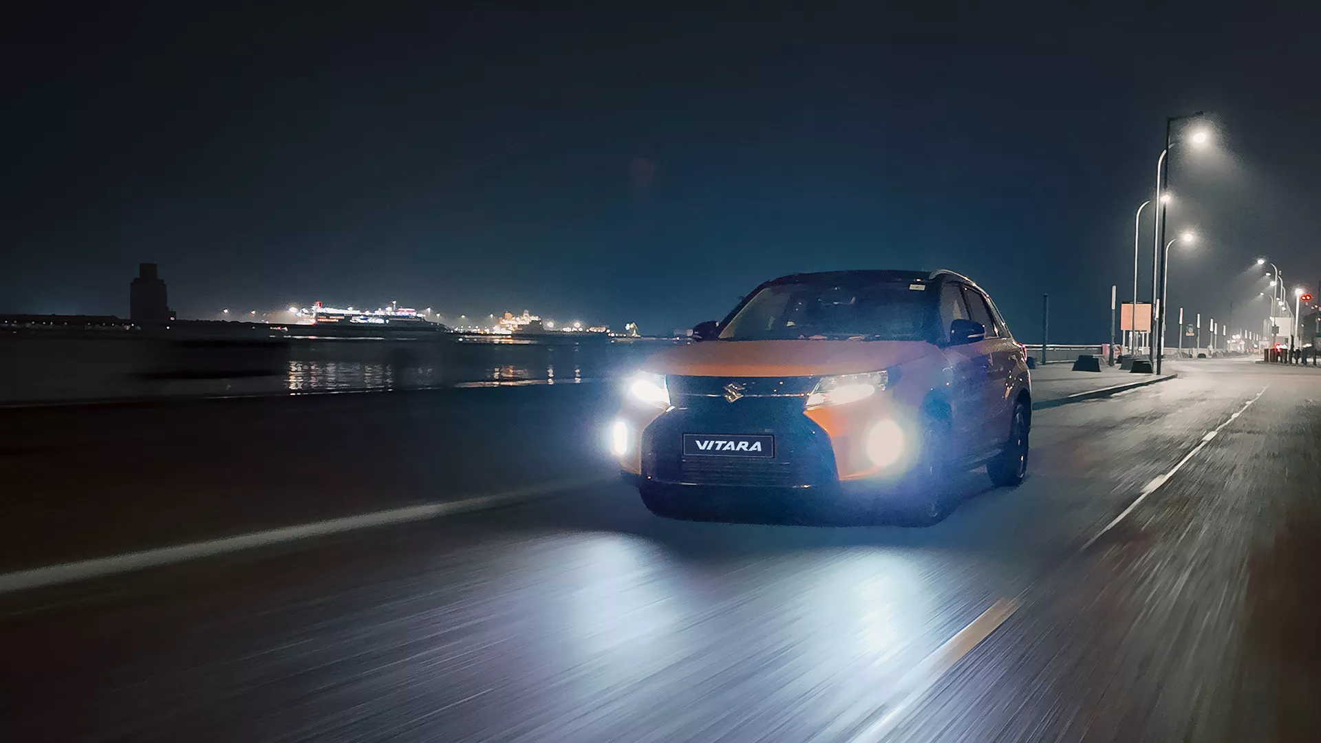 Suzuki Vitara driving along a coastal road at night with headlights on full beam, demonstrating the High Beam Assist feature in low-light conditions.