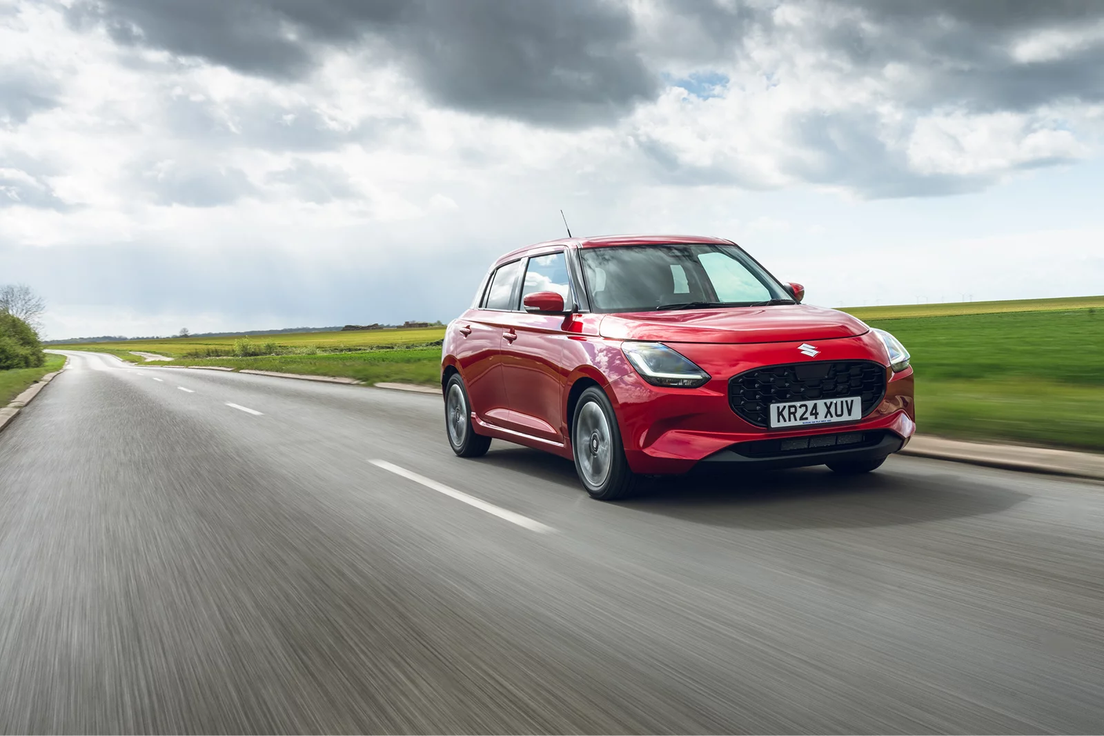 Red Suzuki Swift driving along a countryside road under cloudy skies