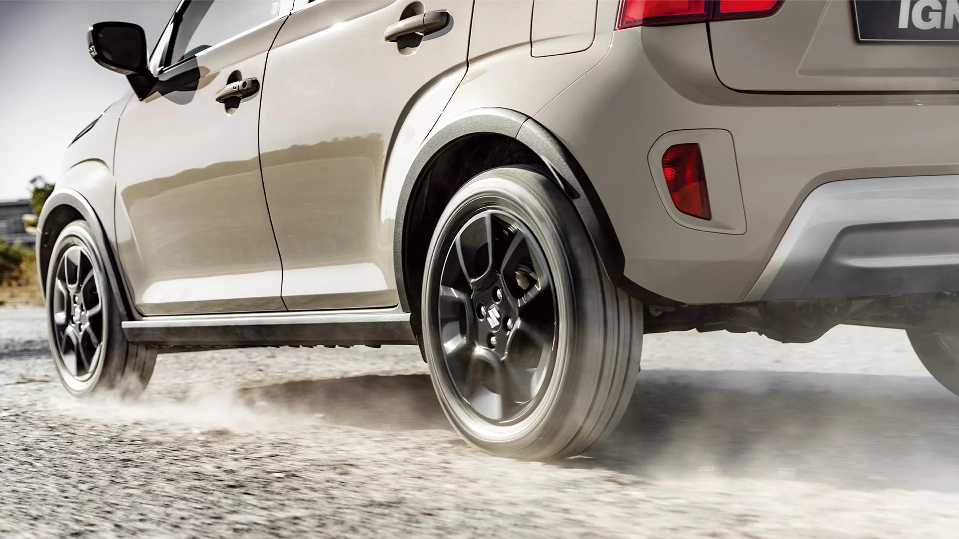 Close-up of Suzuki Ignis in Caravan Ivory driving on gravel, highlighting rear wheel motion and black alloy design.