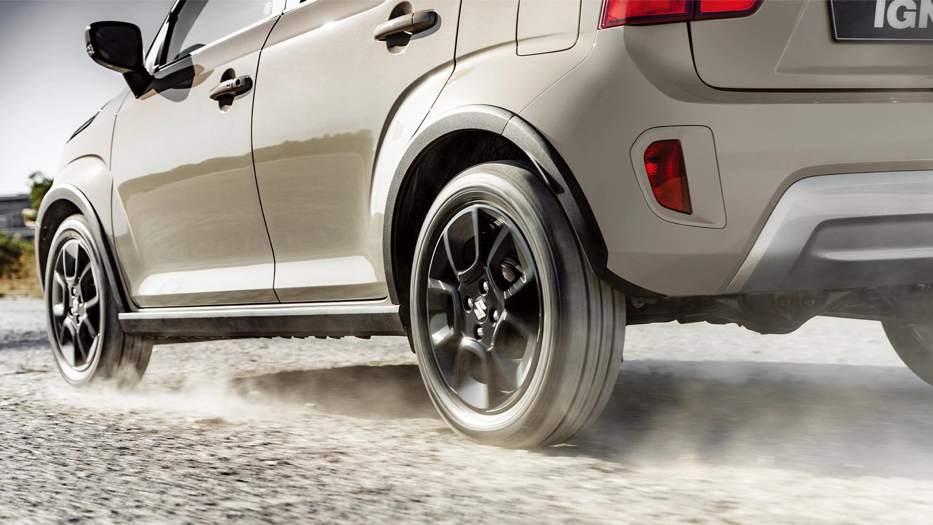Close-up of Suzuki Ignis in Caravan Ivory driving on gravel, highlighting rear wheel motion and black alloy design.