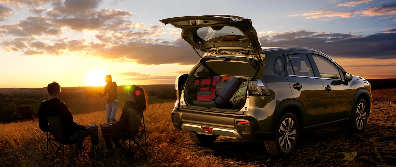 Suzuki e Vitara parked at sunset with open boot packed for a trip, as three people relax outdoors in a countryside setting