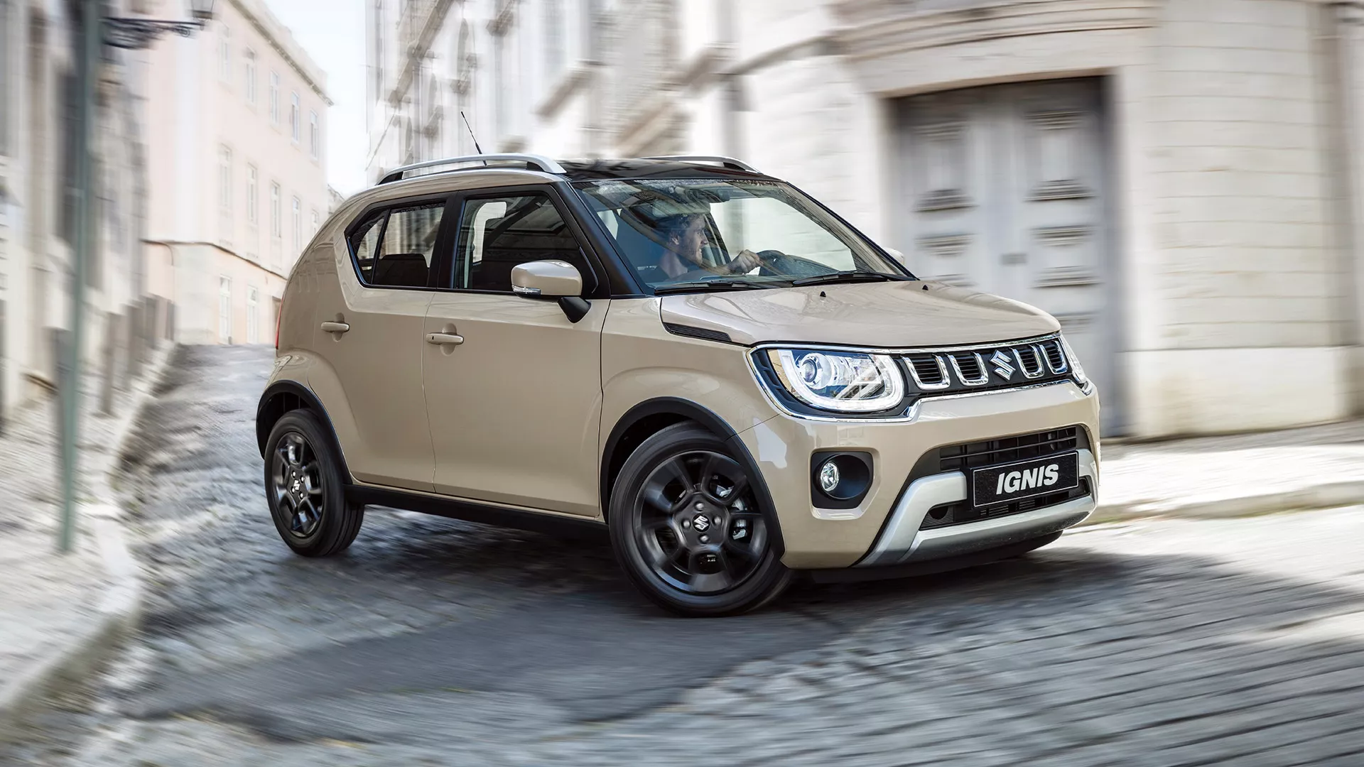 Suzuki Ignis in Caravan Ivory driving through cobbled city street, showing front and side profile with LED headlights and black alloy wheels.