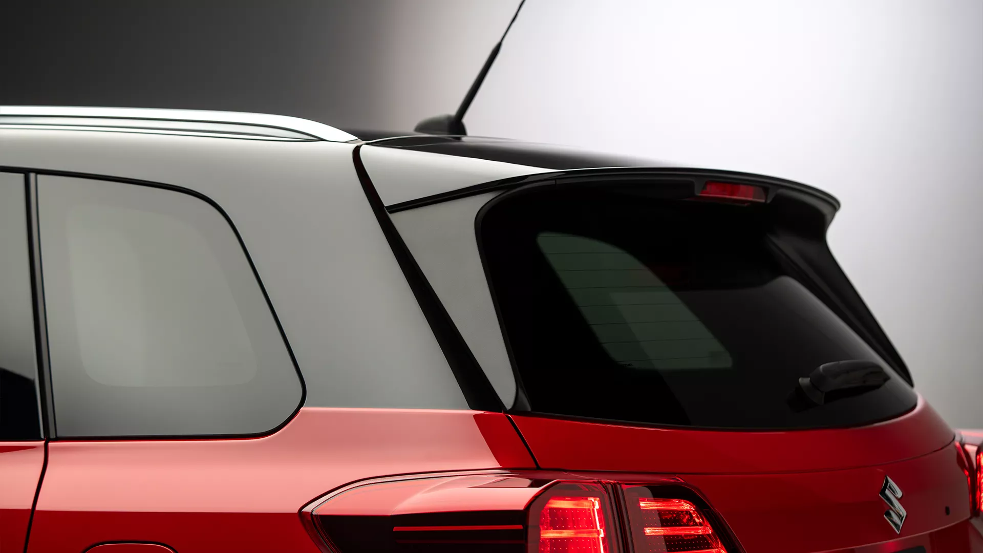 Close-up of the rear upper section of a bright red Suzuki Vitara, highlighting the roof spoiler, cosmic black roof, privacy glass and rear antenna.