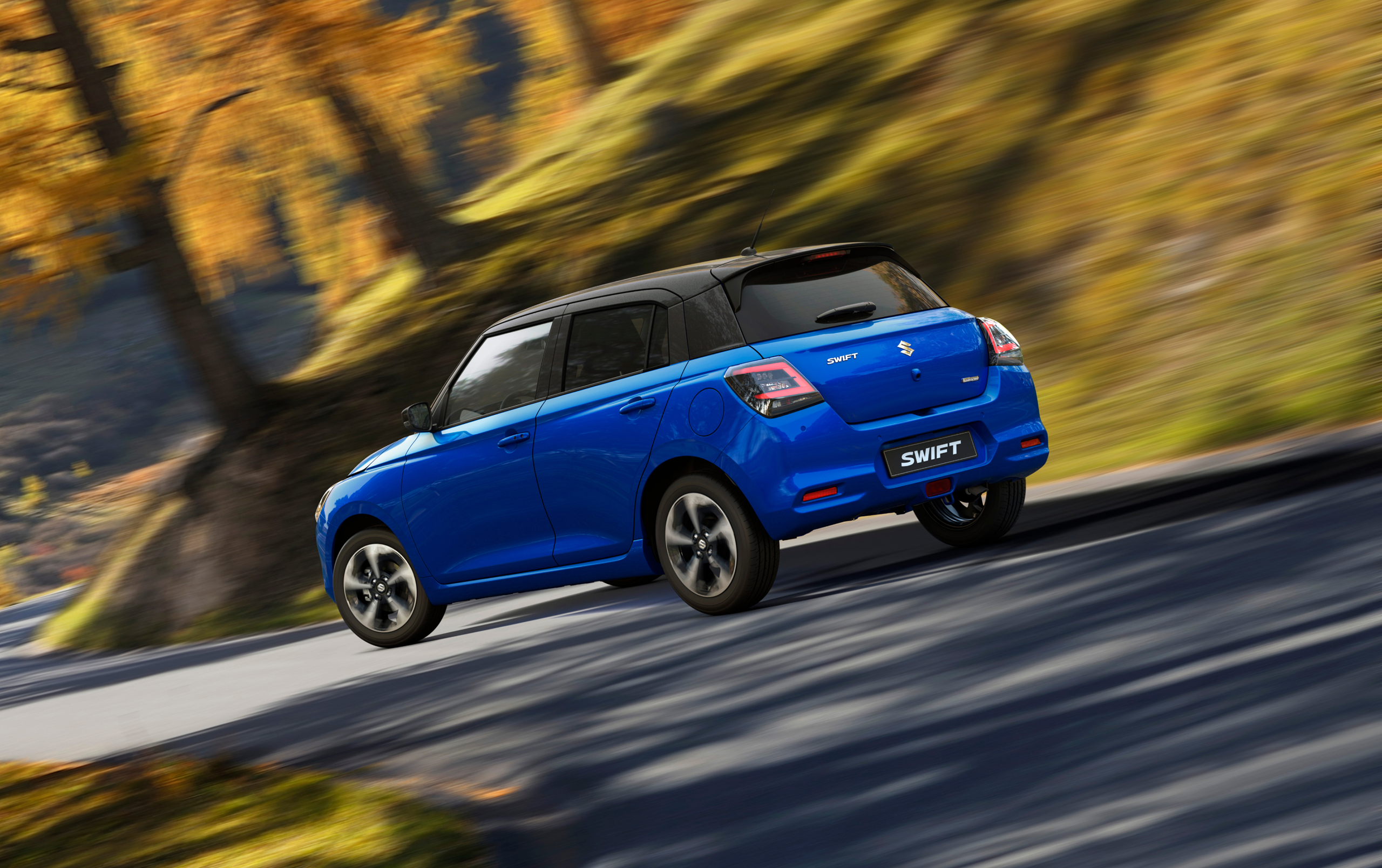 Rear side view of the Suzuki Swift in frontier blue, driving through a winding forest road during autumn with motion blur background.