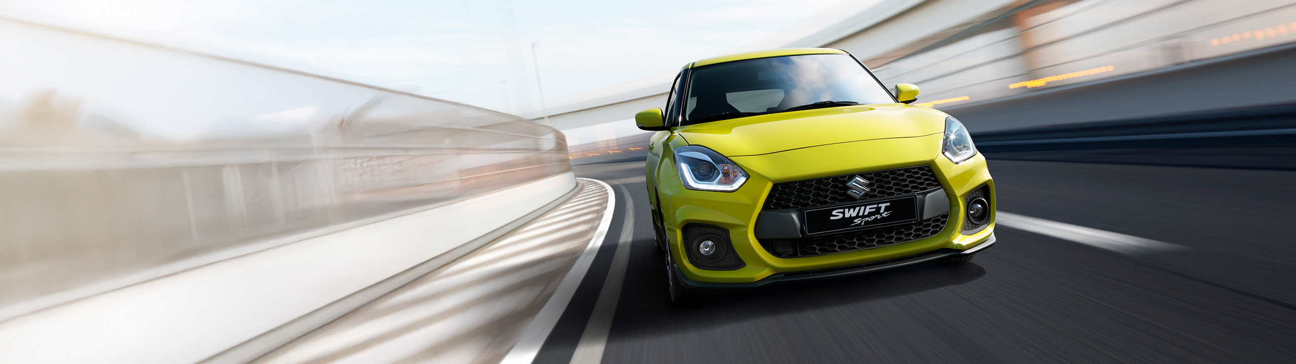 Front view of Suzuki Swift Sport in champion yellow driving at speed on a curved road, highlighting its dynamic performance and sporty design.