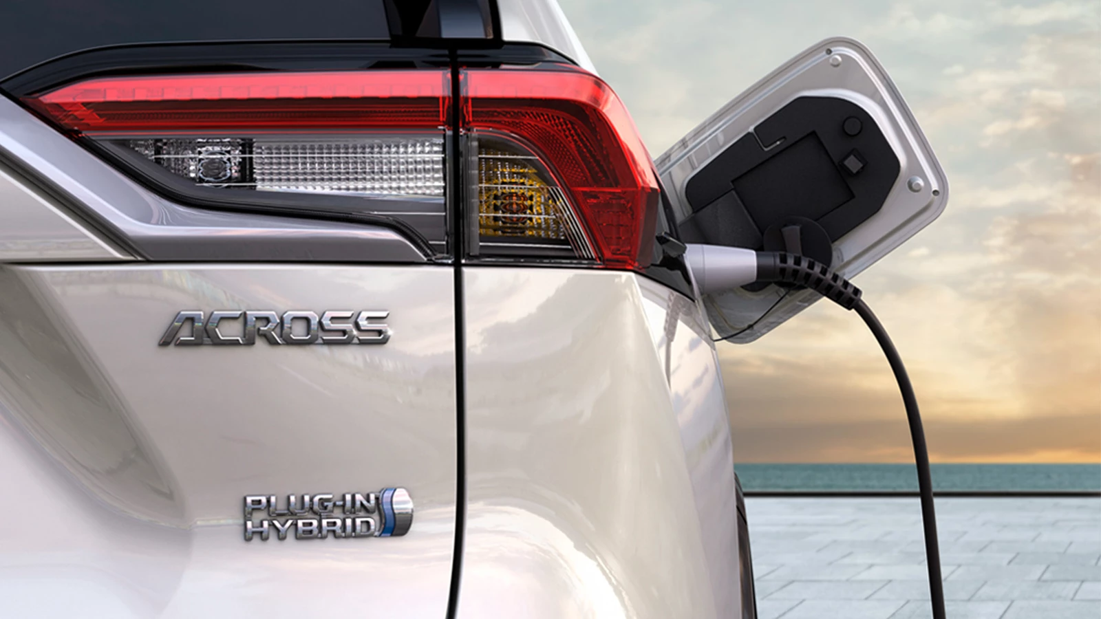 Close-up of the Suzuki Across Plug-in Hybrid charging, showing the rear badge and charging port against a scenic coastal backdrop.