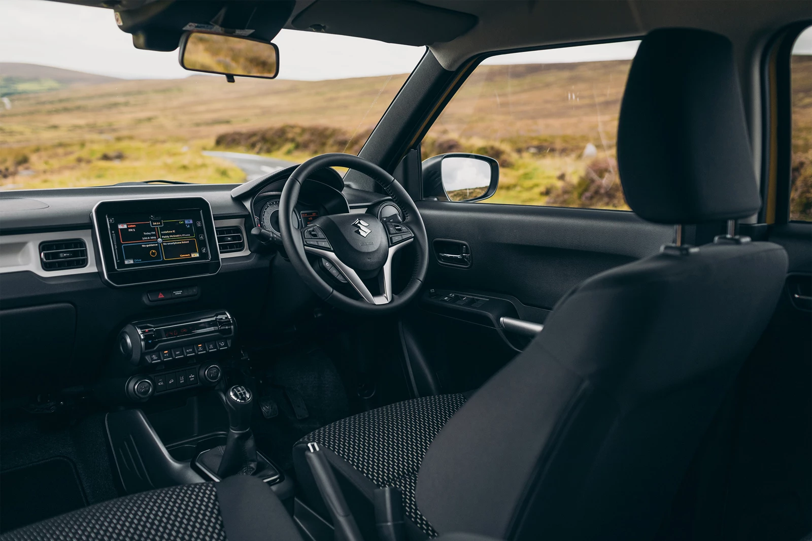 Interior view of Suzuki Igns showcasing dashboard, steering wheel and infotainment screen with ocean view in background.