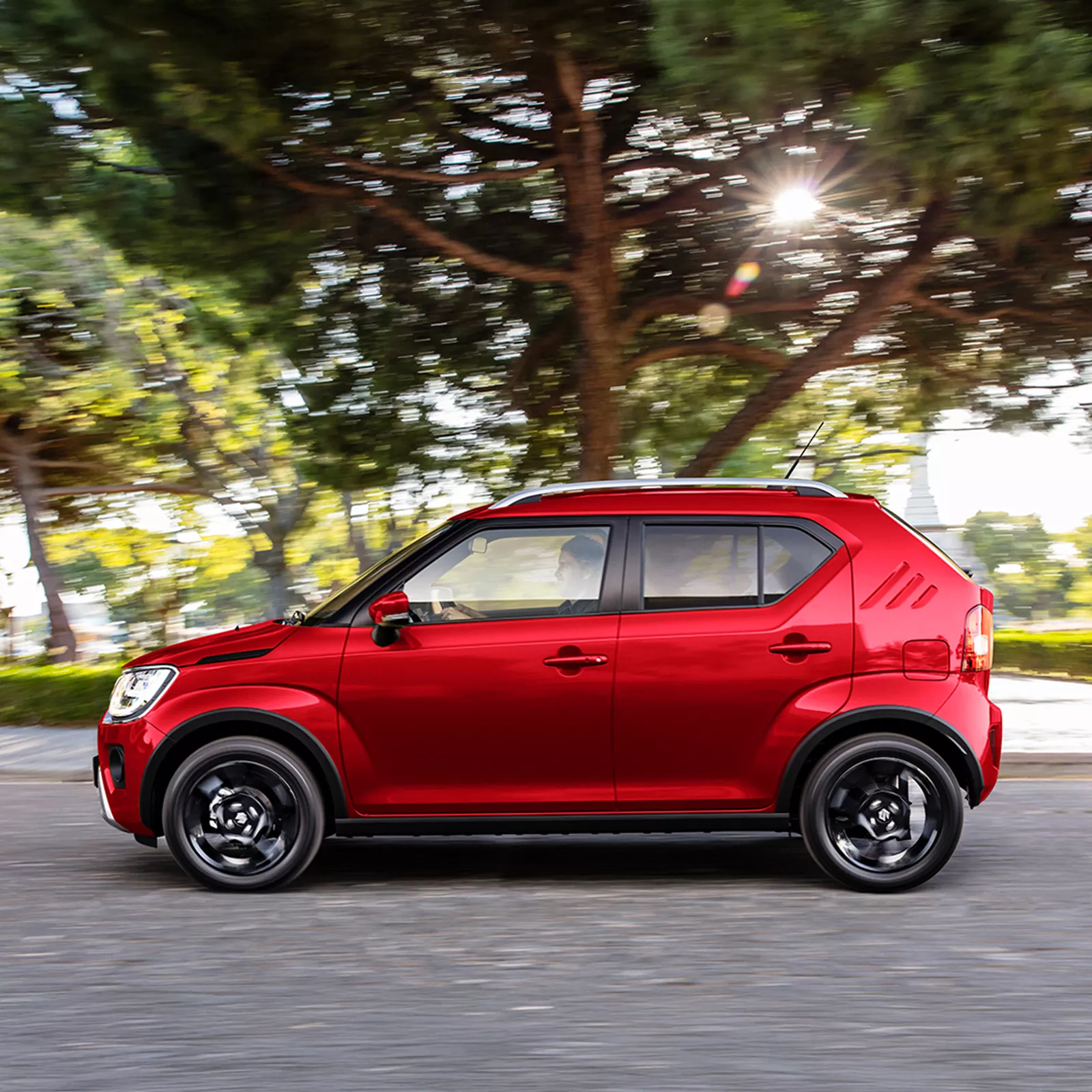 Suzuki Ignis in Burning Red driving through tree-lined street, showcasing side profile with black alloy wheels and roof rails.