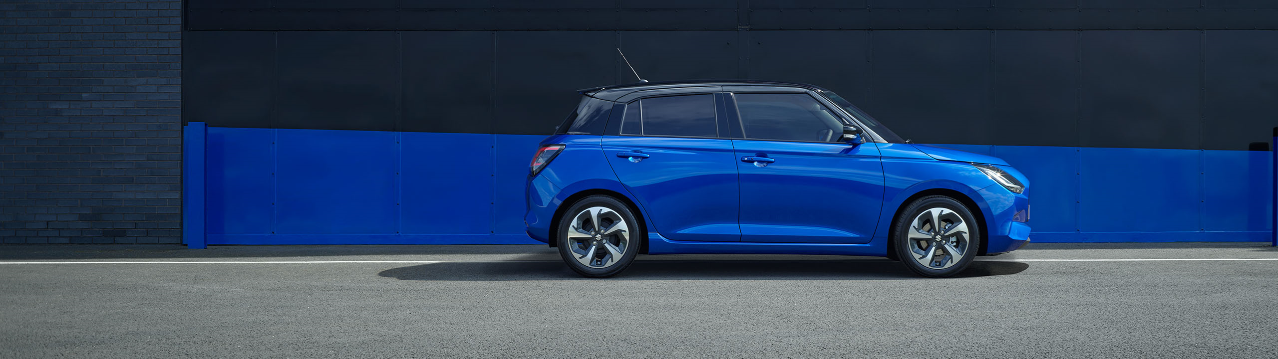 Suzuki Swift in frontier blue pearl metallic with black roof, shown from the side while parked on a street with a blue and black urban backdrop