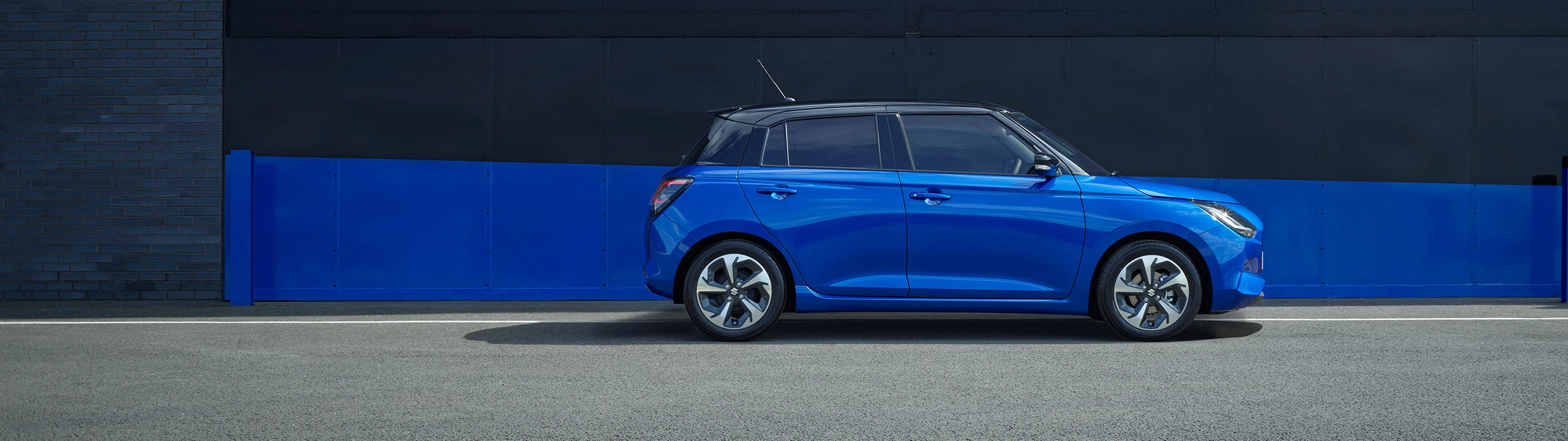 Suzuki Swift in frontier blue pearl metallic with black roof, shown from the side while parked on a street with a blue and black urban backdrop