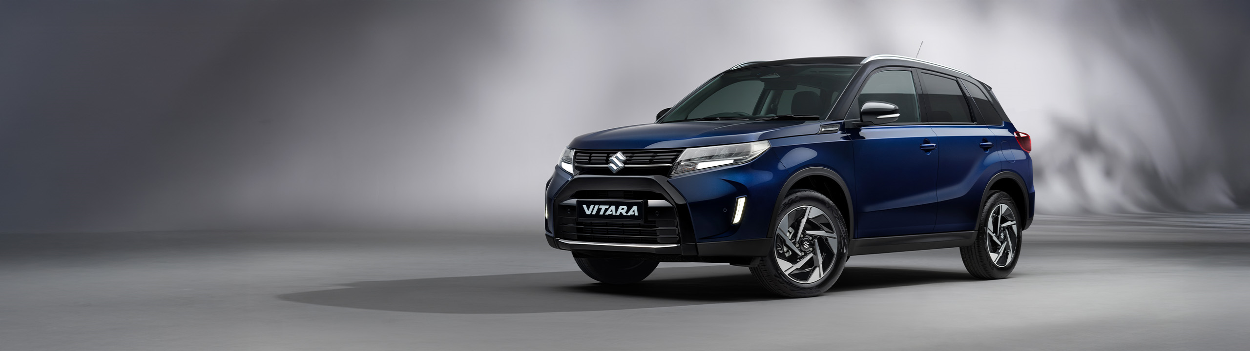 Front three-quarter view of the Suzuki Vitara in sphere blue, displayed in a studio setting with a soft grey background highlighting its bold SUV styling.