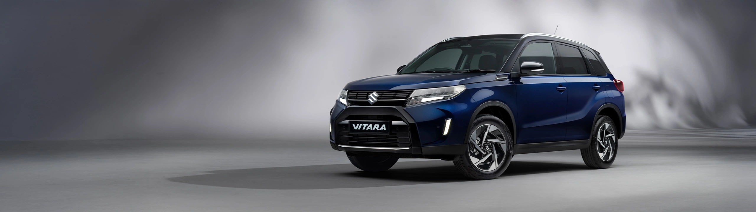 Front three-quarter view of the Suzuki Vitara in sphere blue, displayed in a studio setting with a soft grey background highlighting its bold SUV styling.