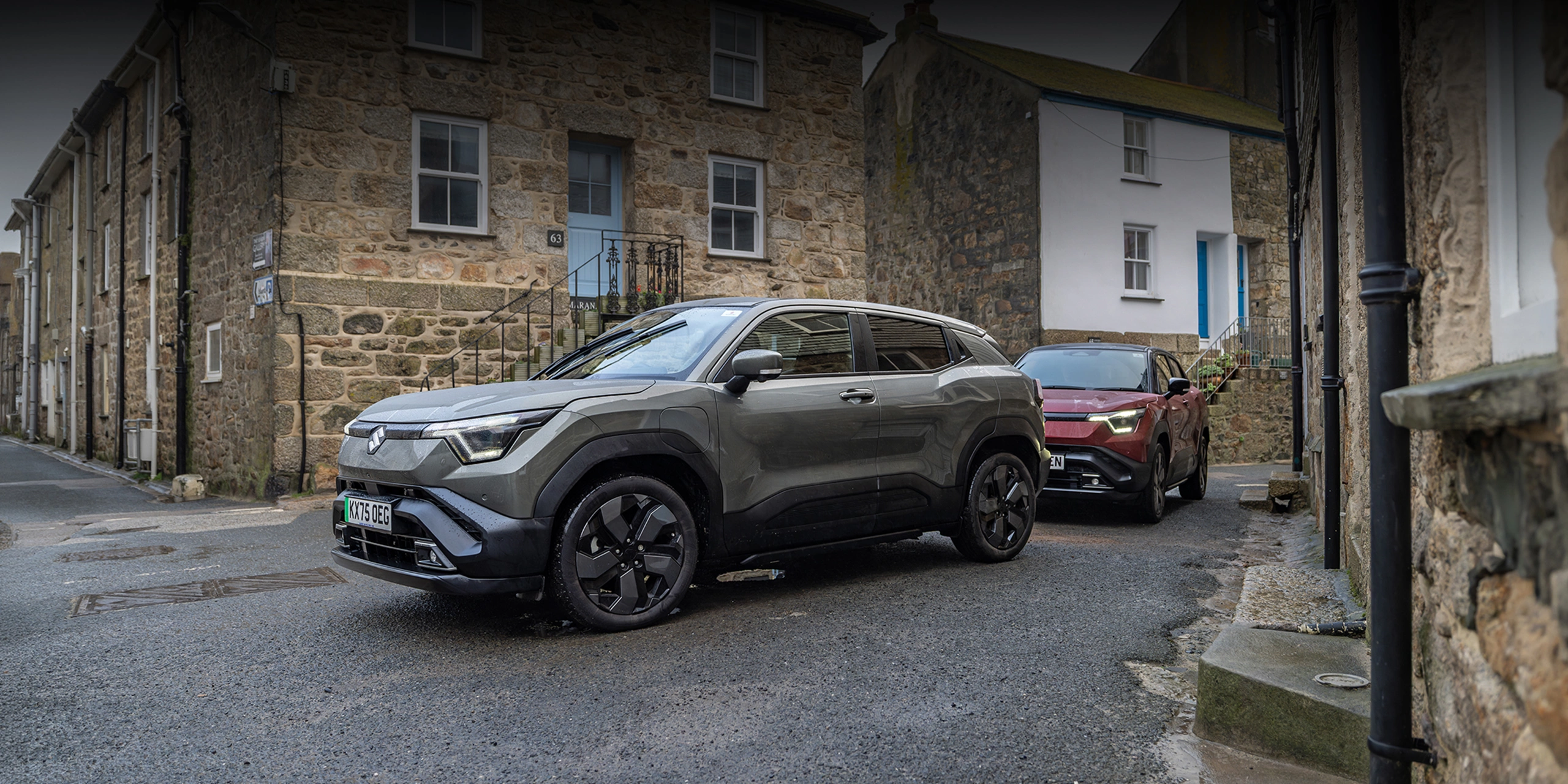 Grey and red Suzuki e Vitara models driving through a village street