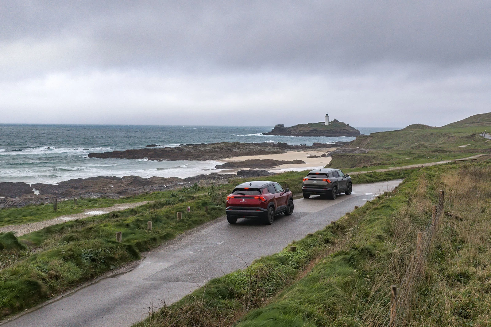 Two Suzuki e Vitara SUVs driving along a coastal road with sea views
