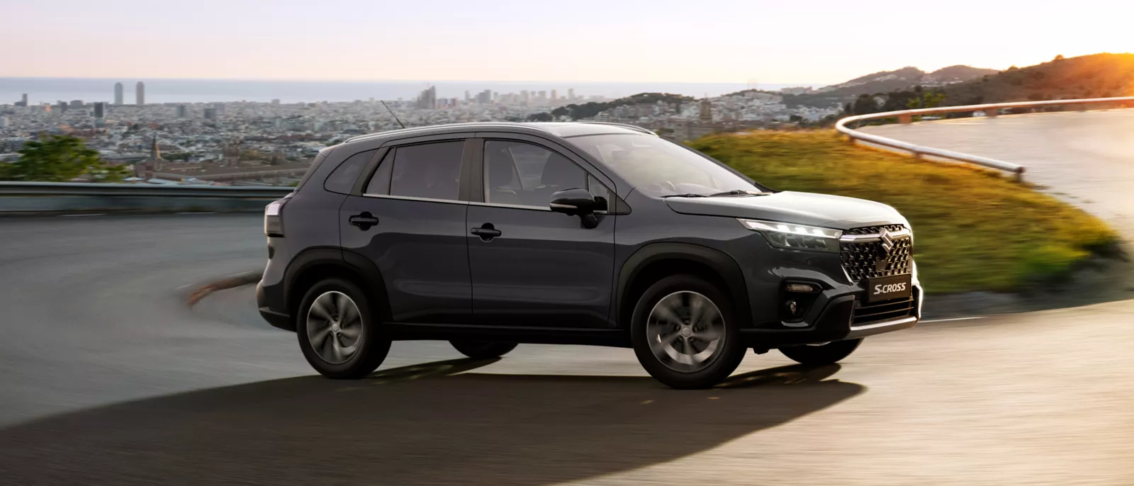 Front and side view of the Suzuki S-Cross in titan dark grey driving on a winding snowy road, showcasing its all-weather capability and rugged design.